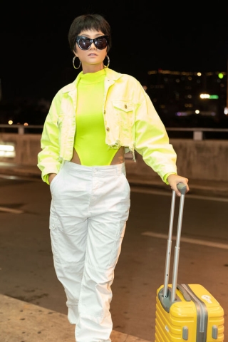 Set đồ thời trang sân bay trẻ trung, năng động của Hoa hậu H'Hen Niê với bodysuit cổ lọ cut - out phần eo mix cùng áo khoác neon xanh sáng lóa phối cùng quần trắng